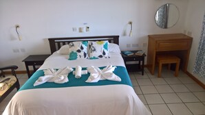 Una cama bien hecha con sábanas blancas y una colcha color turquesa, una mesita de noche con libros, una mesita auxiliar de madera, un espejo en la pared y un pequeño banco de madera.