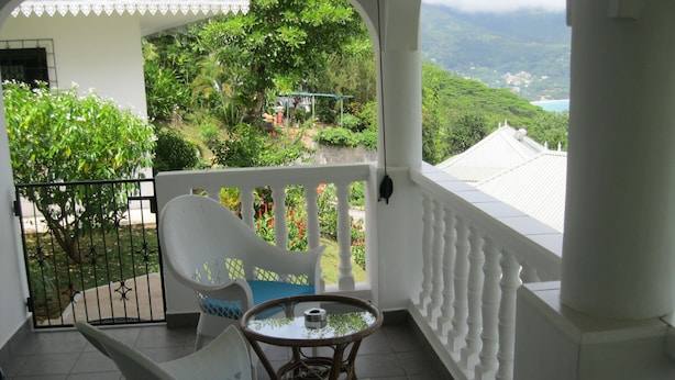 Un balcón con una silla blanca y una pequeña mesa, con vistas a un exuberante jardín y un paisaje montañoso.