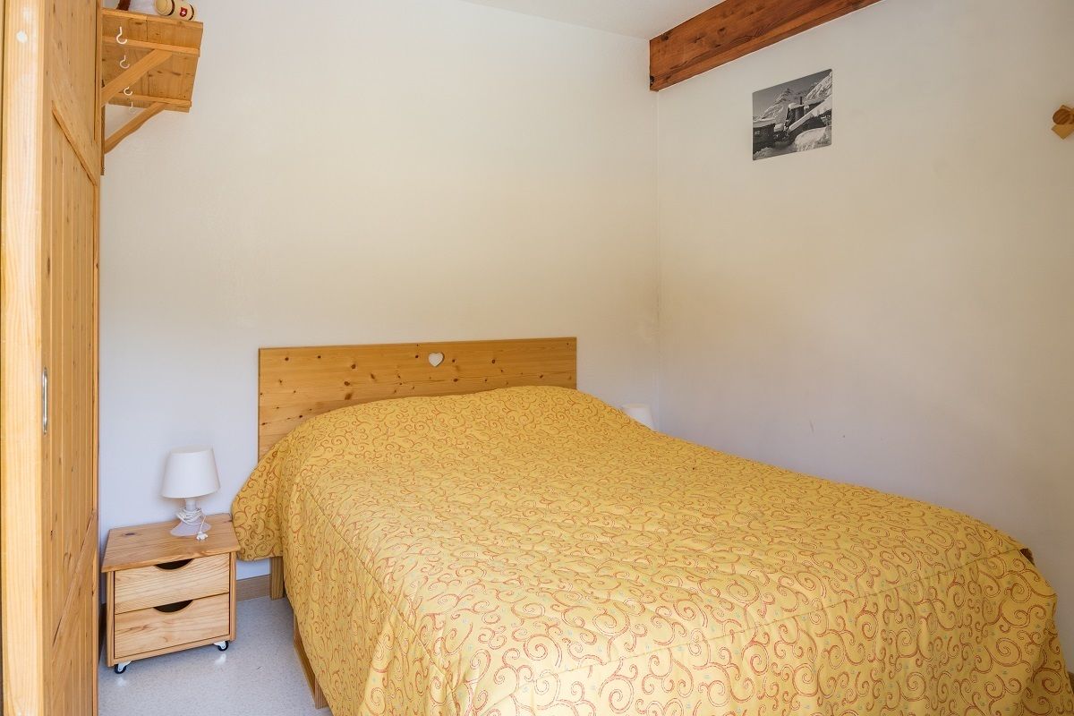 A bedroom with a wooden headboard, a yellow patterned bedspread, a bedside table with a lamp, and a small drawer unit.