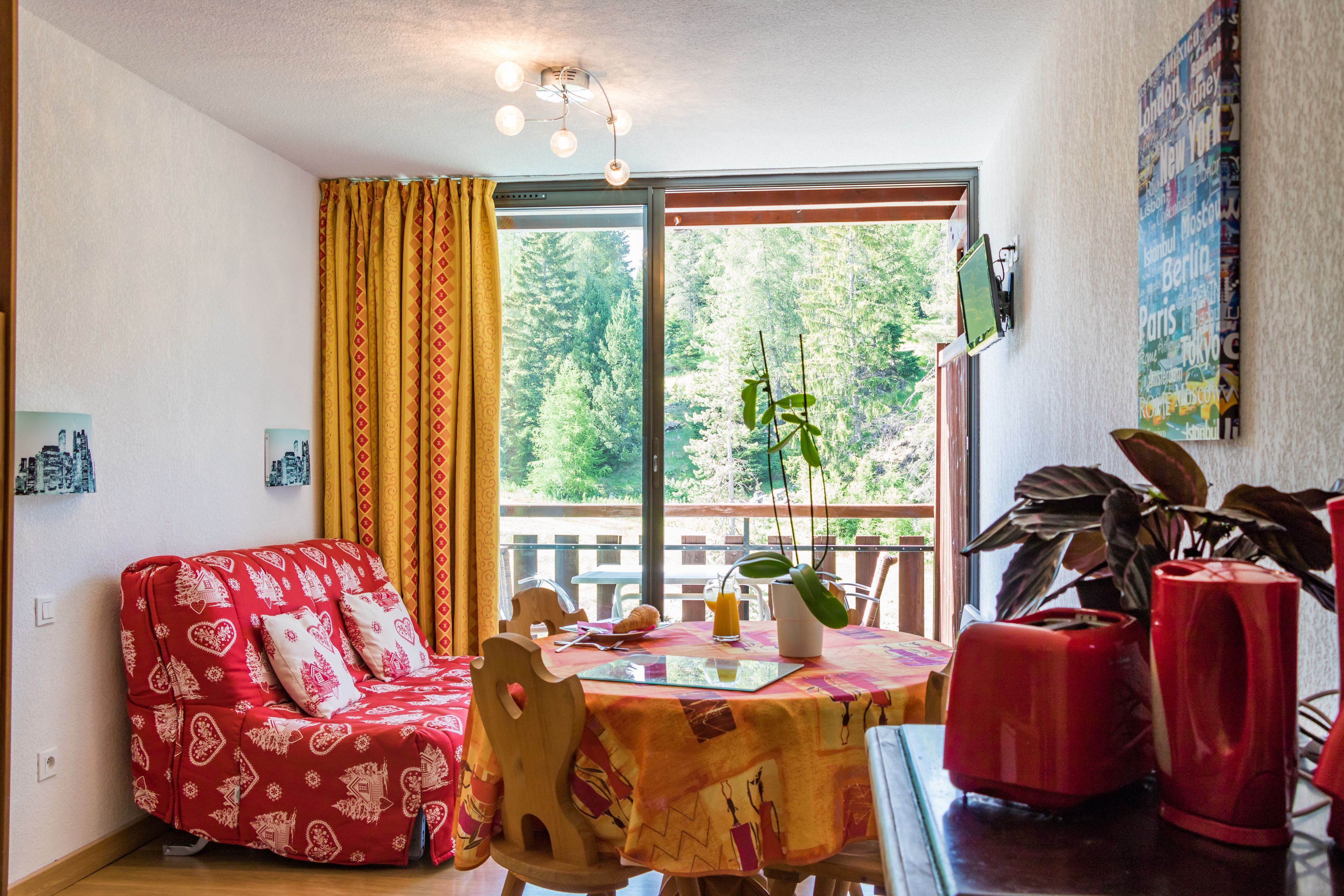 A cozy living room with a red patterned sofa, a wooden dining table with a red tablecloth, and a large window offering a view of trees.