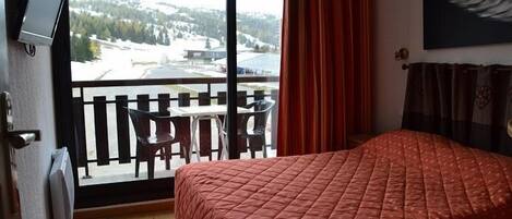 A hotel room with a bed, a TV mounted on the wall, a balcony with a table and chairs, and a view of a snowy landscape.