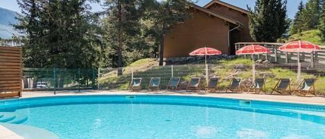 A swimming pool with lounge chairs and umbrellas, surrounded by a wooden fence and trees.