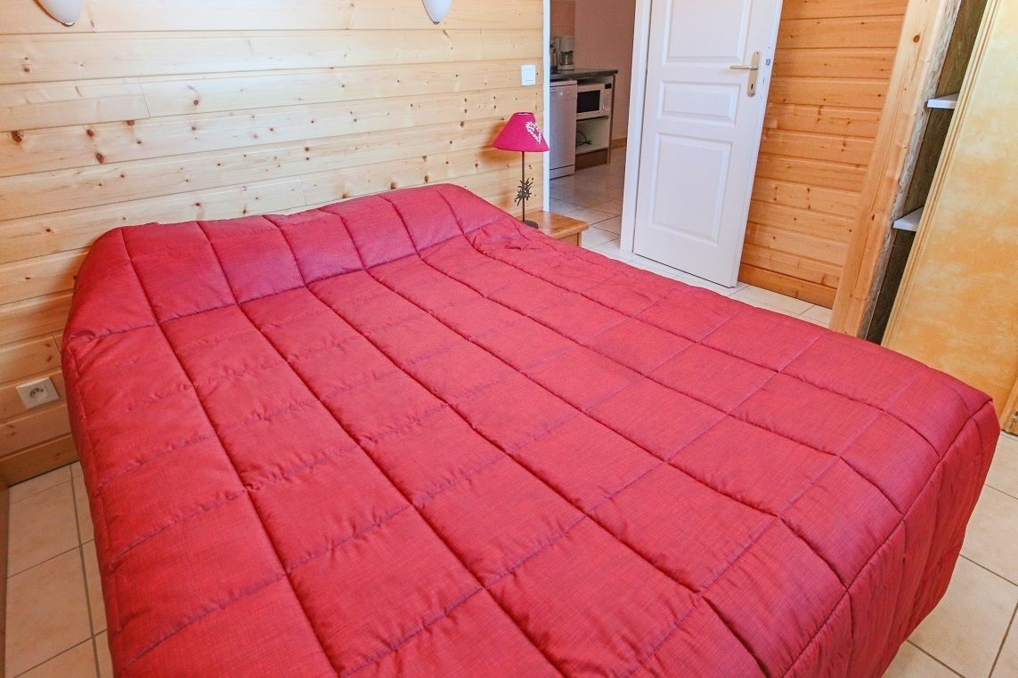 Una habitación con una cama cubierta por un edredón rojo, paredes de madera, una mesita con una lámpara rosa y una puerta que da a una zona de cocina.