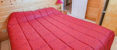 Una habitación con una cama cubierta por un edredón rojo, paredes de madera, una mesita con una lámpara rosa y una puerta que da a una zona de cocina.