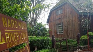 Un letrero de madera con el texto 'NARIBU RIVER LODGE' y 'NARIBU NATURAL HOSPITAL', rodeado de exuberante follaje y un edificio rústico.