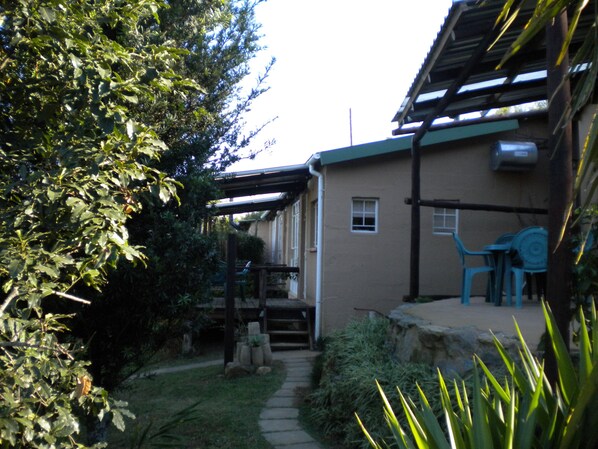 Un edificio residencial con un patio cubierto, un sendero de piedra que lleva a la entrada y un pequeño jardín con diversas plantas.