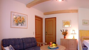 A cozy living room with a blue patterned sofa, a round wooden table with a fruit bowl, and a cabinet with a lamp. - Ferienwohnung Taubensee, 1-2 Personen, 45 qm, Bad + Dusche/WC, Südbalkon, TV, 1 Schlafzimmer, Nichtraucher (Ramsau bei Berchtesgaden)