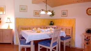A dining area with a wooden table set for four, surrounded by chairs with blue cushions. A white cabinet with a lace doily is to the left, and a clock is on the wall to the right. - Ferienwohnung Taubensee, 1-2 Personen, 45 qm, Bad + Dusche/WC, Südbalkon, TV, 1 Schlafzimmer, Nichtraucher (Ramsau bei Berchtesgaden)