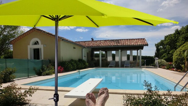 A swimming pool with a sun umbrella, a covered patio, and a house with a tiled roof. - Gîtes La Bakénia (Villeneuve-sur-Lot)