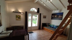A cozy room with a wooden staircase, a sofa with a colorful blanket, a dining table with chairs, a radiator, and a large window with a view of the garden. - Gîtes La Bakénia (Villeneuve-sur-Lot)