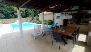 A covered outdoor area with a pool, a dining table with chairs, and a kitchen area. - Gîtes La Bakénia (Villeneuve-sur-Lot)