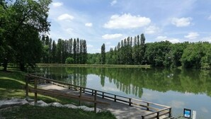 A wooden bridge over a calm lake, surrounded by lush greenery and trees. - Gîtes La Bakénia (Villeneuve-sur-Lot)