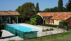 A swimming pool with a tiled deck, surrounded by a fence, and adjacent to a building with a tiled roof. - Gîtes La Bakénia (Villeneuve-sur-Lot)