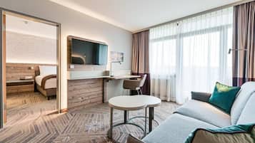 A modern hotel room with a sofa, a small table, a TV mounted on the wall, and a large window with curtains.