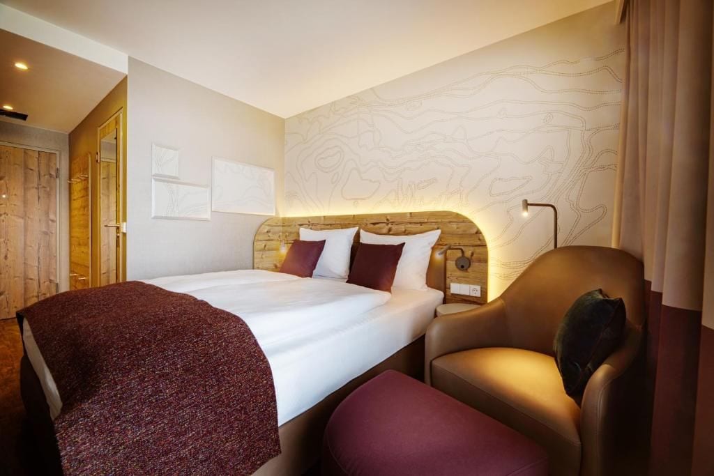 Una habitación de hotel con una cama grande, cabecera de madera, zona de estar con una silla y reposapiés, y una pared con un motivo decorativo.