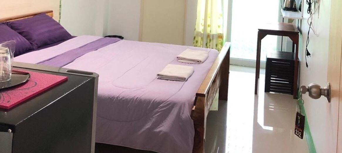 A neatly made bed with a purple bedspread and two folded towels, a bedside table with a glass and a pink mat, and a wooden nightstand.