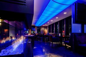 A modern interior with a blue-lit ceiling, a fire feature, and a seating area with tables and chairs. - Hotel Stern by EMPA (Ulm)