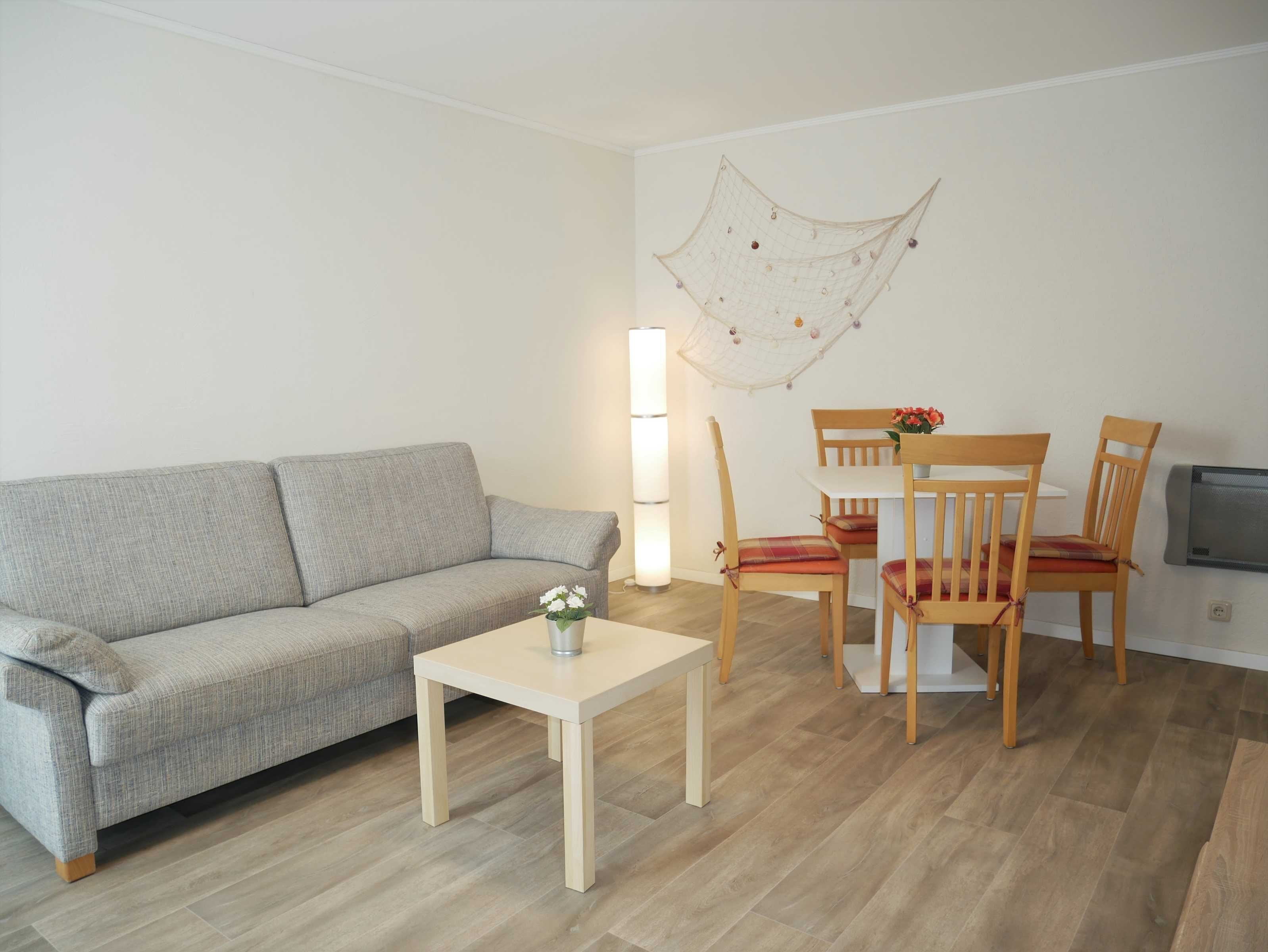 A living room with a grey sofa, a wooden dining table with chairs, a small white table, and a decorative wall hanging.