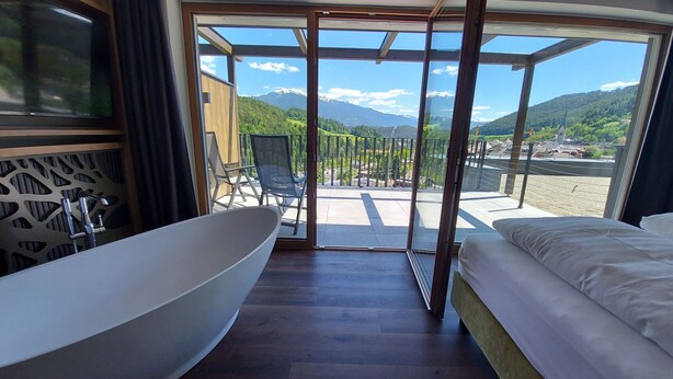 A modern bathroom with a large bathtub, a shower, and a glass-enclosed balcony with a view of mountains and a village. - Molaris Lodge (Rio di Pusteria)