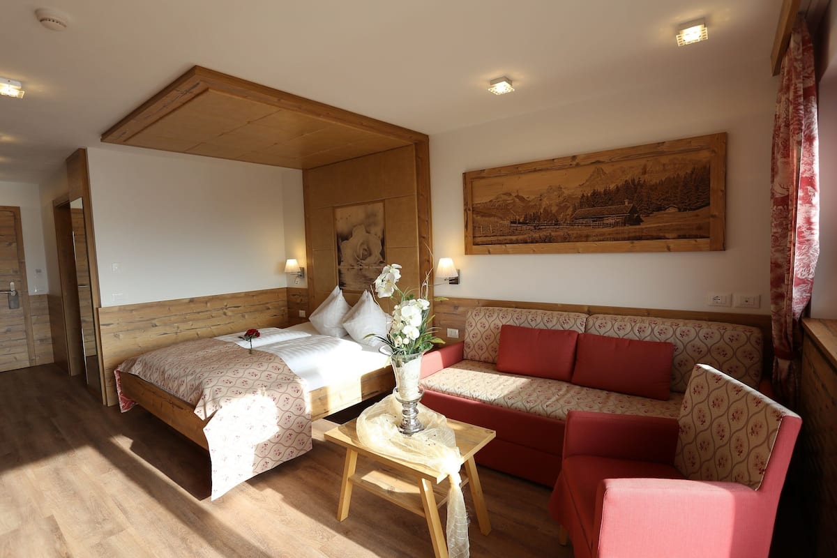 Une chambre d’hôtel avec un plafond en bois, un lit recouvert d’une couvre-lit à motifs floraux, un canapé avec des coussins rouges, une petite table sur laquelle est posé un vase de fleurs, et un tableau accroché au mur.
