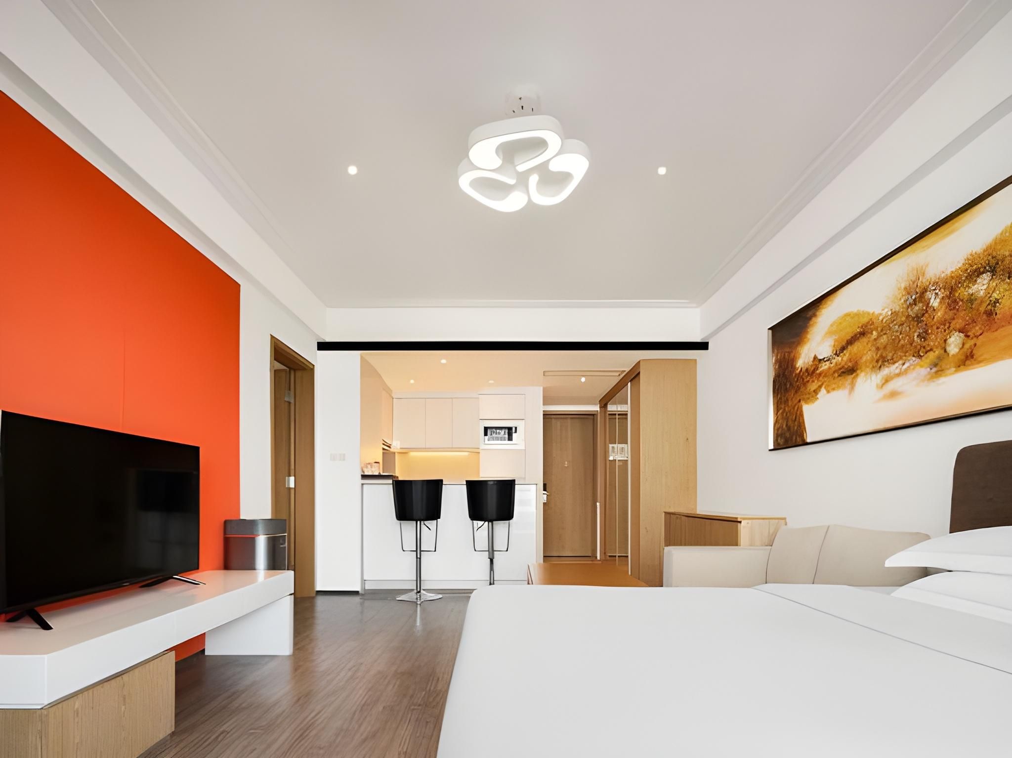 A modern hotel room with a large bed, a TV, a dining area, and a kitchen in the background.