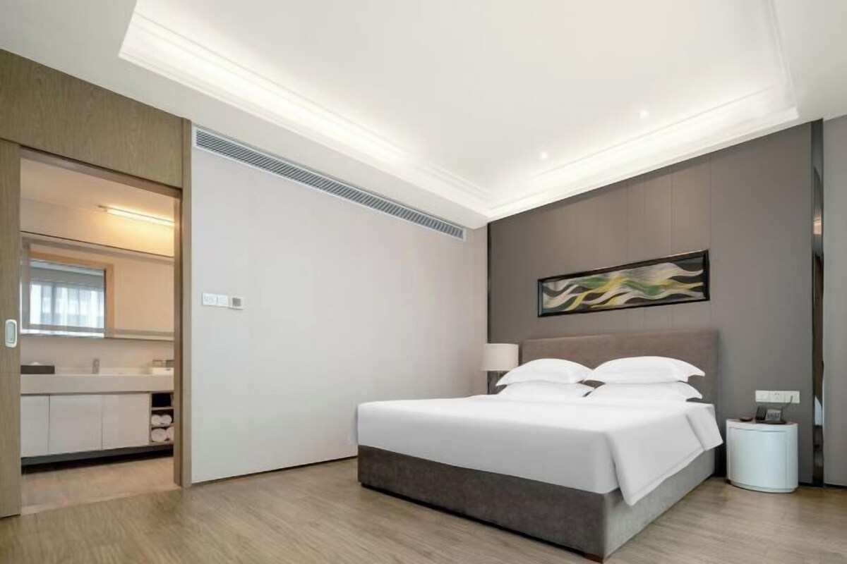 A modern hotel room with a bed, bedside table, lamp, and a painting on the wall.