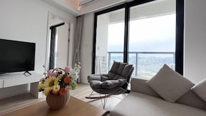 A modern living room with a flat-screen TV, a grey rocking chair, a sofa, and a large window with a city view. - Dahengqin Superior Talent Hotel (Hengqin Port Branch) (Zhuhai)