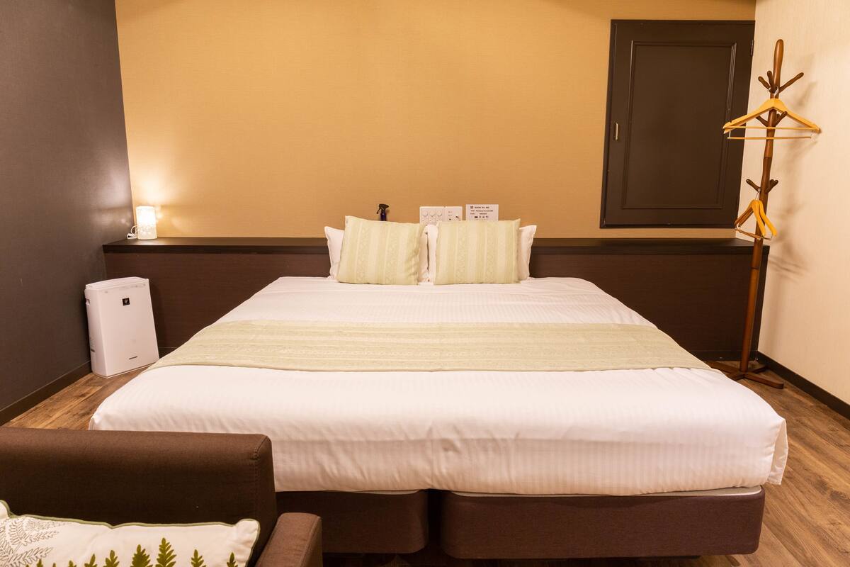 A hotel room with a large bed, two pillows, a bedside table, a lamp, and a coat rack.