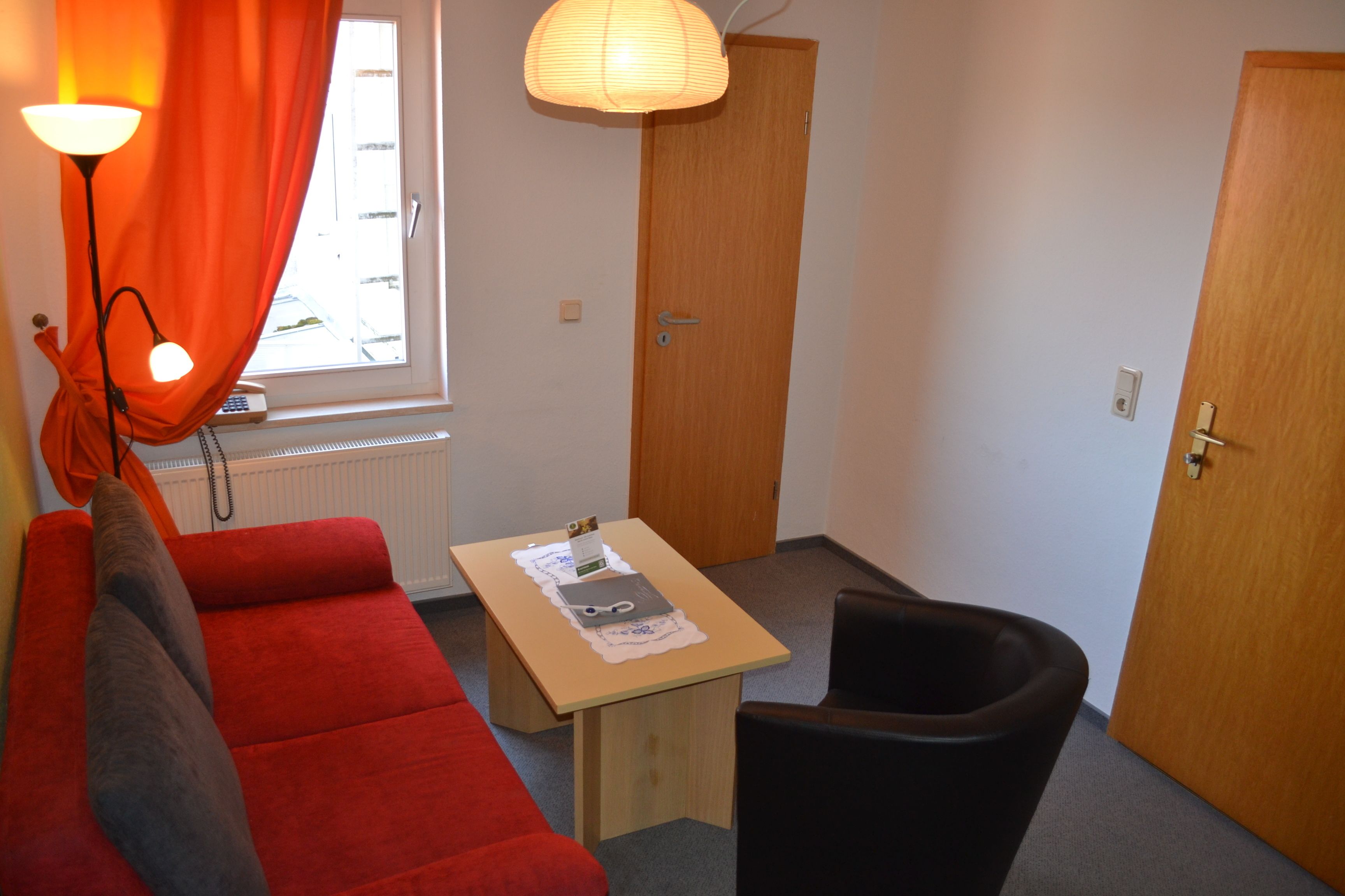 A small room with a red sofa, a wooden table with a lamp, a black chair, and a wooden door.