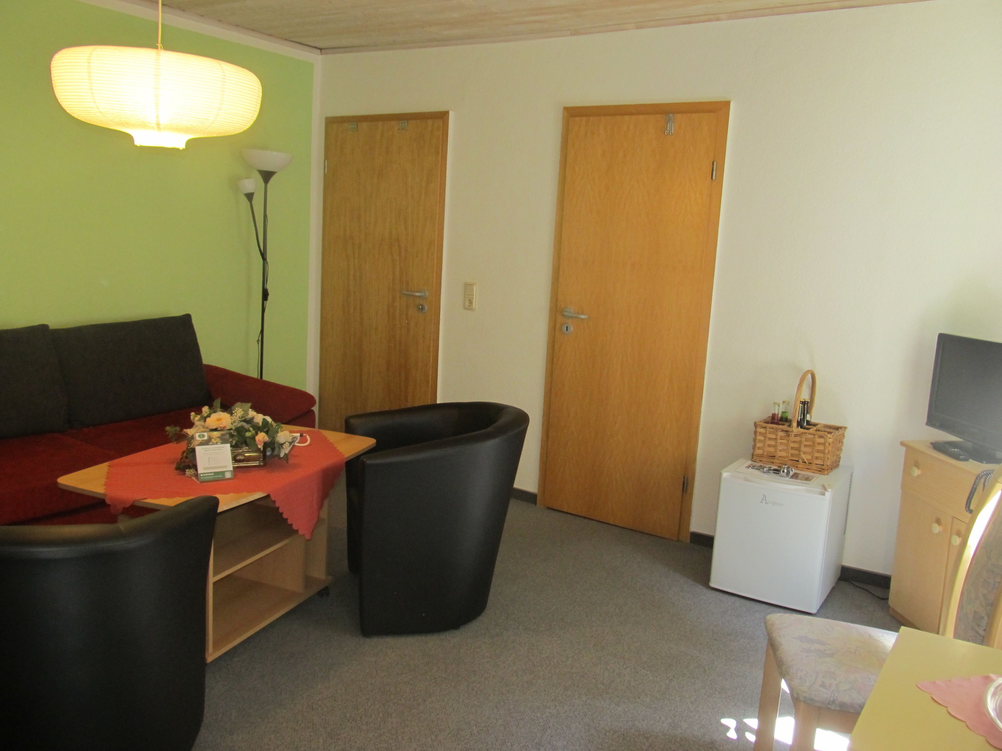 Ein Zimmer mit einer Couch, zwei Stühlen, einem kleinen Tisch mit roter Tischdecke, einer Lampe, einem Fernseher und einem Schrank mit einem Korb.