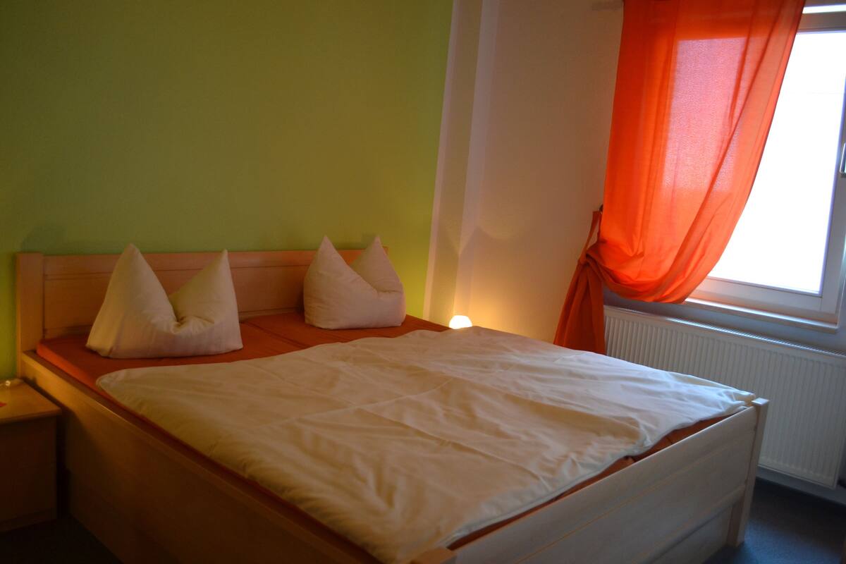 Un lit avec un matelas et des oreillers blancs, une tête de lit en bois, une table de chevet et une fenêtre avec des rideaux orange.