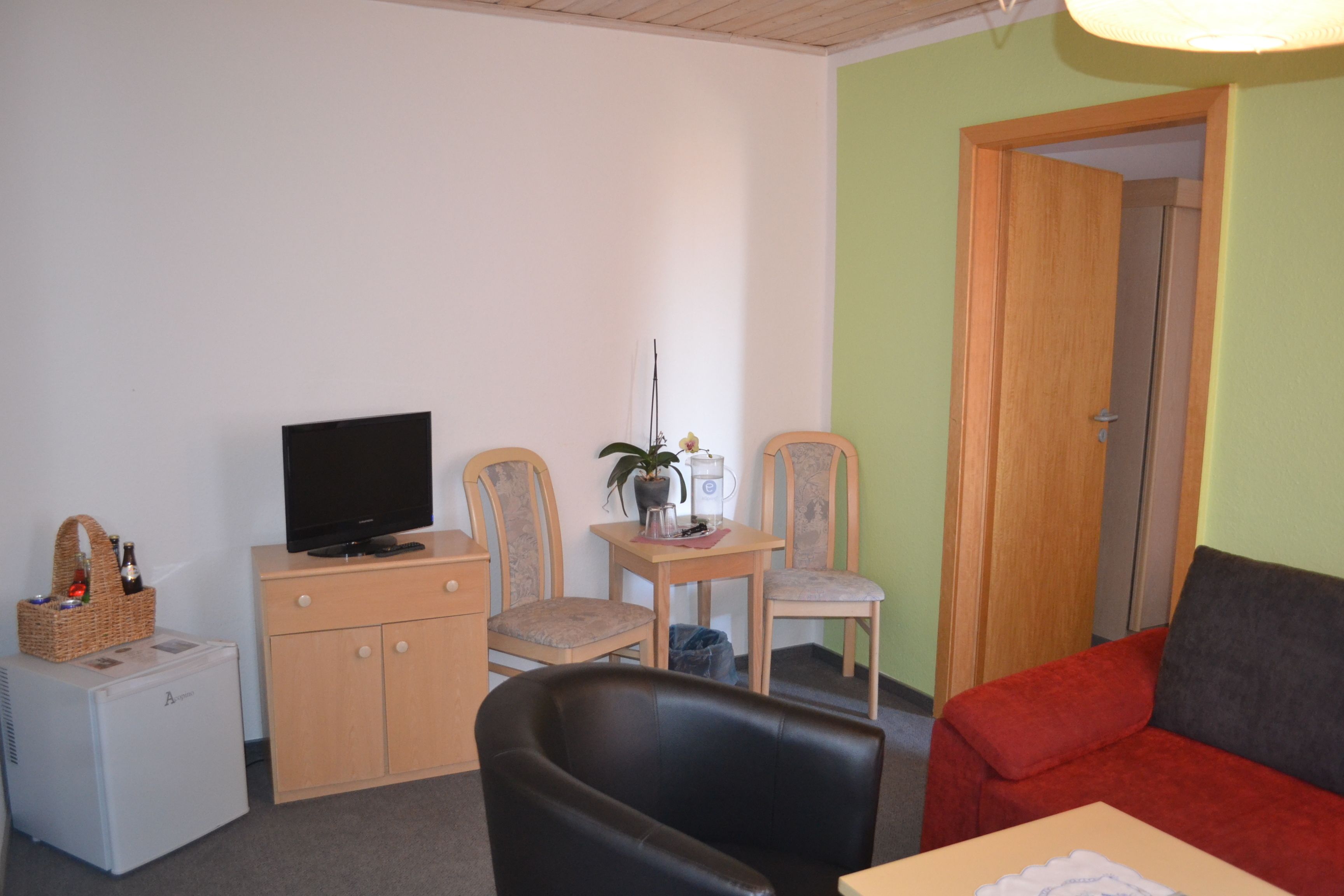 Ein kompakter Wohnraum mit Fernseher auf einem Schrank, zwei Stühle, einem kleinen Tisch mit Pflanze, einem roten Sofa und einer Tür zu einem anderen Raum.