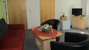 Un salon avec un canapé rouge, un fauteuil noir, une table basse en bois sur laquelle est posé un vase de fleurs, un téléviseur sur un meuble en bois et une lampe.