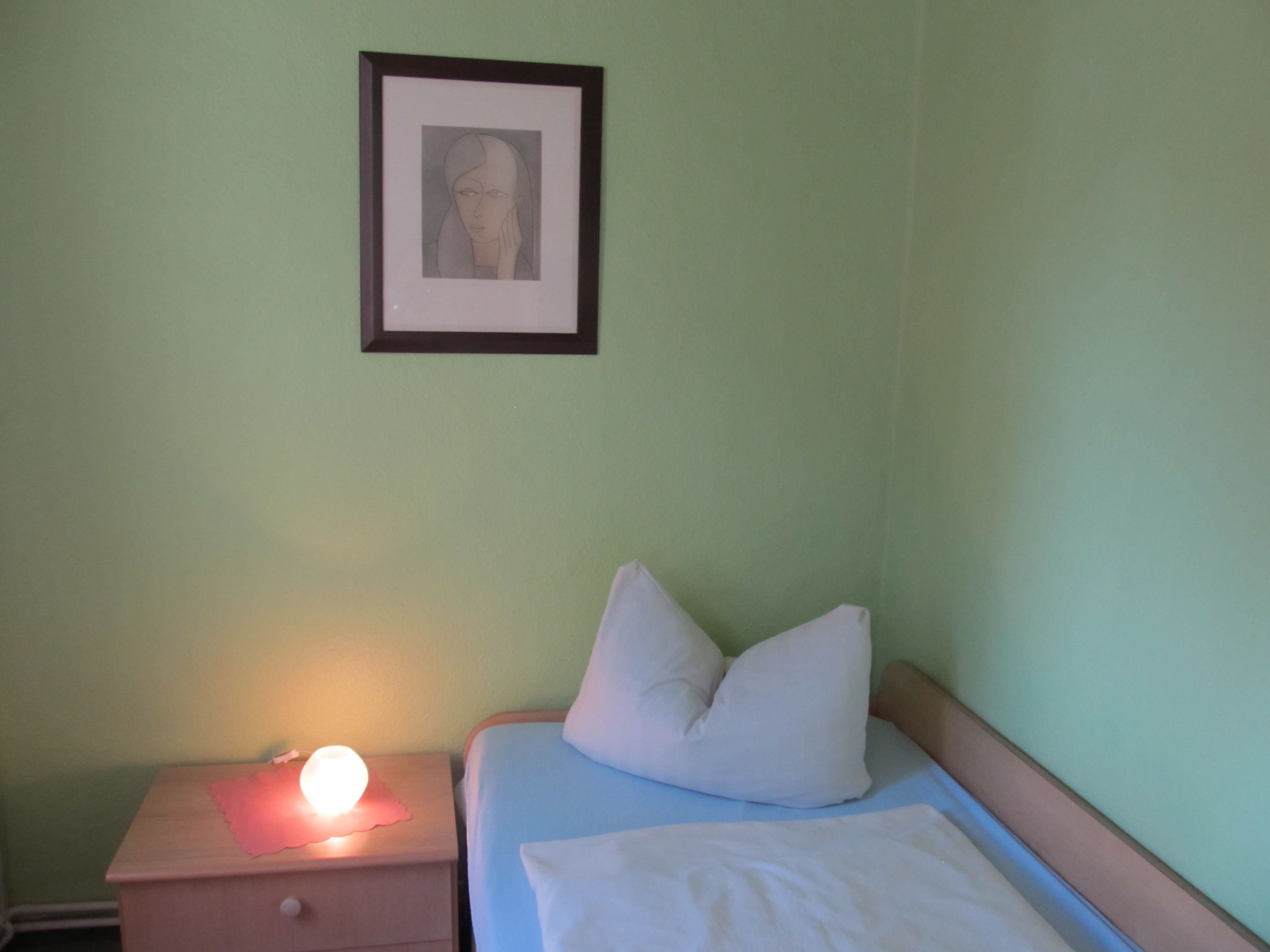 Un dormitorio con cama, almohadas, una mesita de noche con lámpara y un cuadro en la pared.