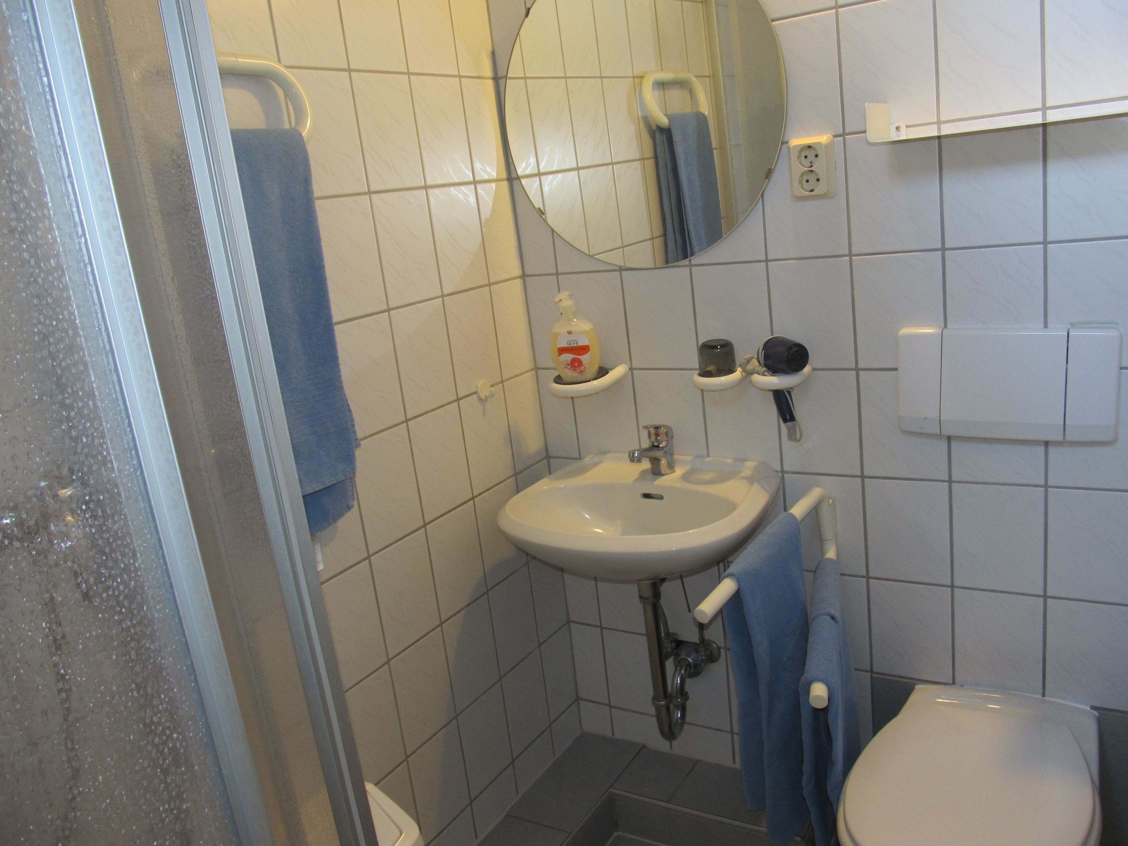 Un baño con lavamanos blanco, espejo e inodoro. Hay una toalla azul colgada en la pared y un dispensador de jabón en el lavamanos.