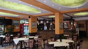 A restaurant with multiple tables and chairs, a Coca-Cola refrigerator, a bar area with a stone wall, and ceiling lights. - Tanya Hotel (Sunny Beach)