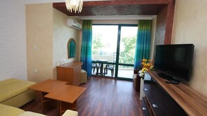 A modern living room with a flat-screen TV, wooden furniture, a dining area with a table and chairs, and a balcony with a view. - Tanya Hotel (Sunny Beach)
