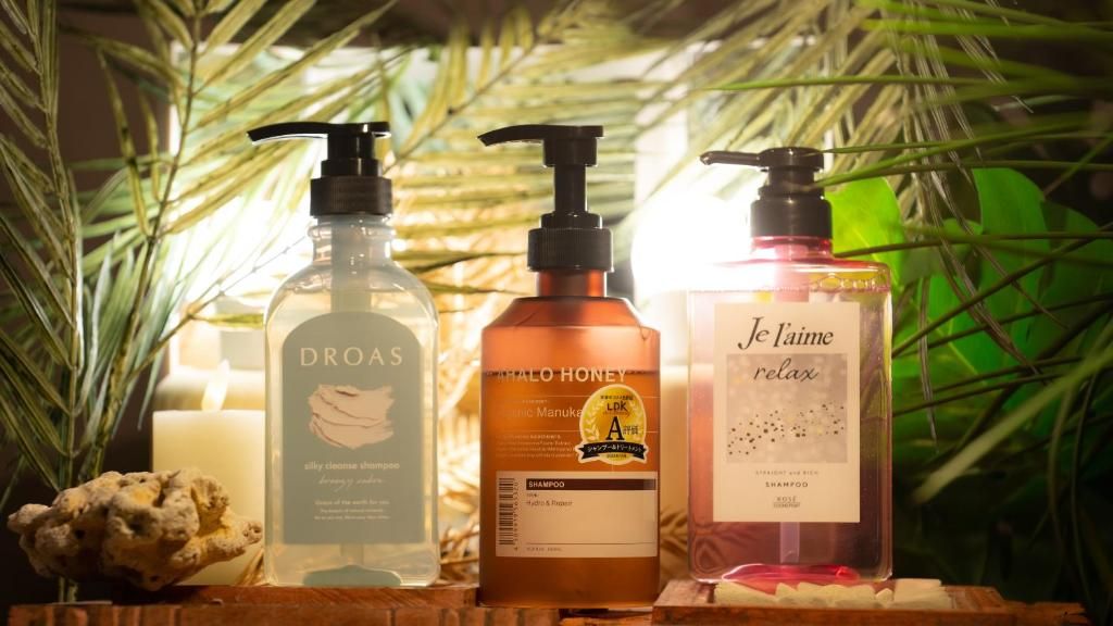 Three bottles of shampoo with pumps, placed on a wooden surface with a candle and a rock.