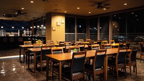 A spacious dining area with multiple wooden tables and chairs, a bar area with stools, and large windows providing a view of the cityscape at night. - Rycom Crystal Hotel (Okinawa)