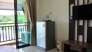 A hotel room with a bed, a small refrigerator, a TV, a window with curtains, and a balcony with a view of greenery.