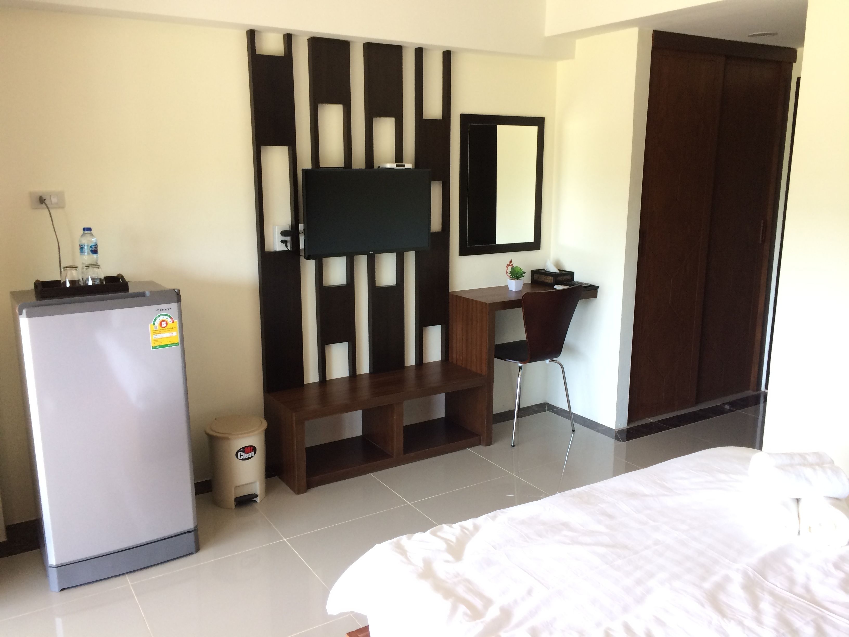 A hotel room with a bed, a desk with a chair, a television mounted on a wall, a wardrobe, and a refrigerator.