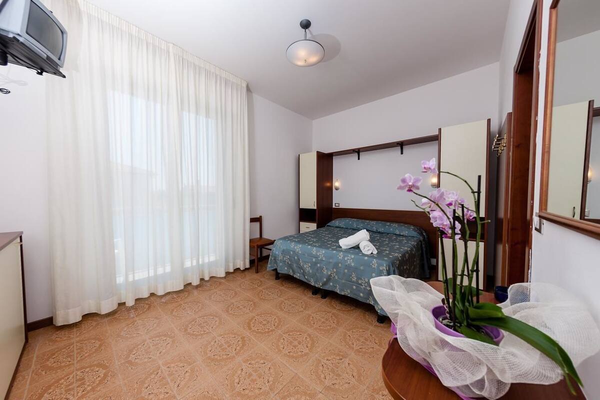 A hotel room with a bed, a TV, a window with curtains, a mirror, and a potted orchid.