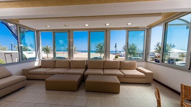 A spacious living area with a large beige sectional sofa, two ottomans, and a wicker chair. The room features large windows offering a view of a beach and cityscape. - Hotel Sayonara (Senigallia)