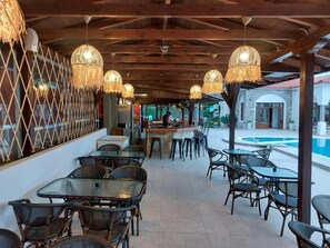 Un espace extérieur aménagé avec des tables et des chaises en osier, des lampes en osier suspendues au-dessus, et une piscine en arrière-plan.