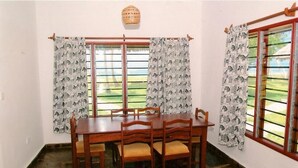 Comedor con mesa y sillas de madera, dos ventanas con cortinas estampadas y una lámpara colgante.