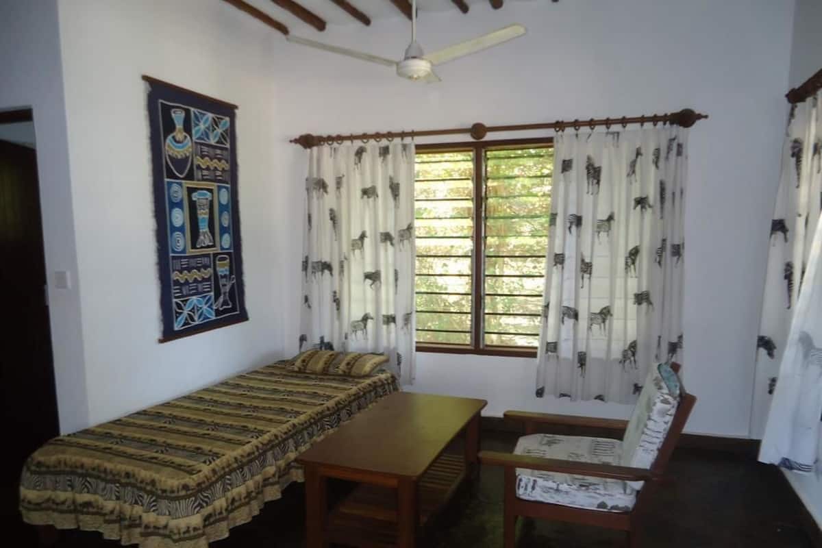 A room with a bed, a wooden table, a chair, a ceiling fan, and a window with patterned curtains.