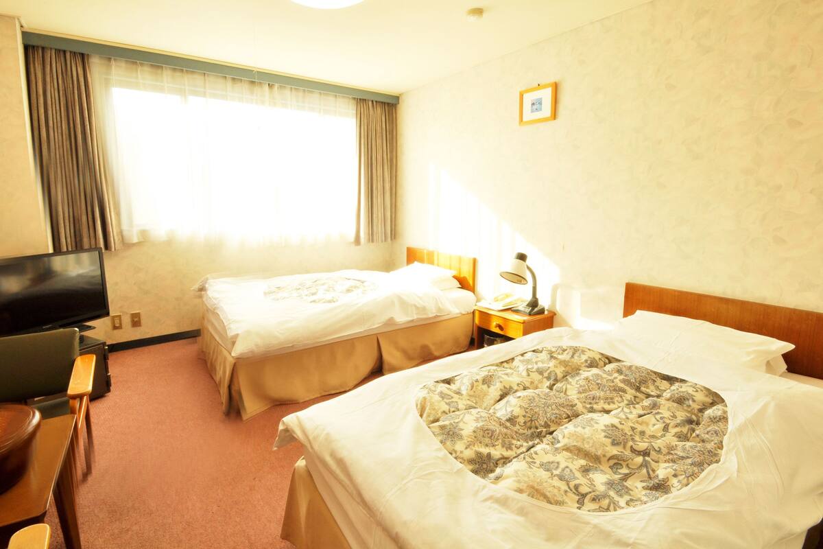 A hotel room with two beds, a desk, a TV, and a window with curtains.