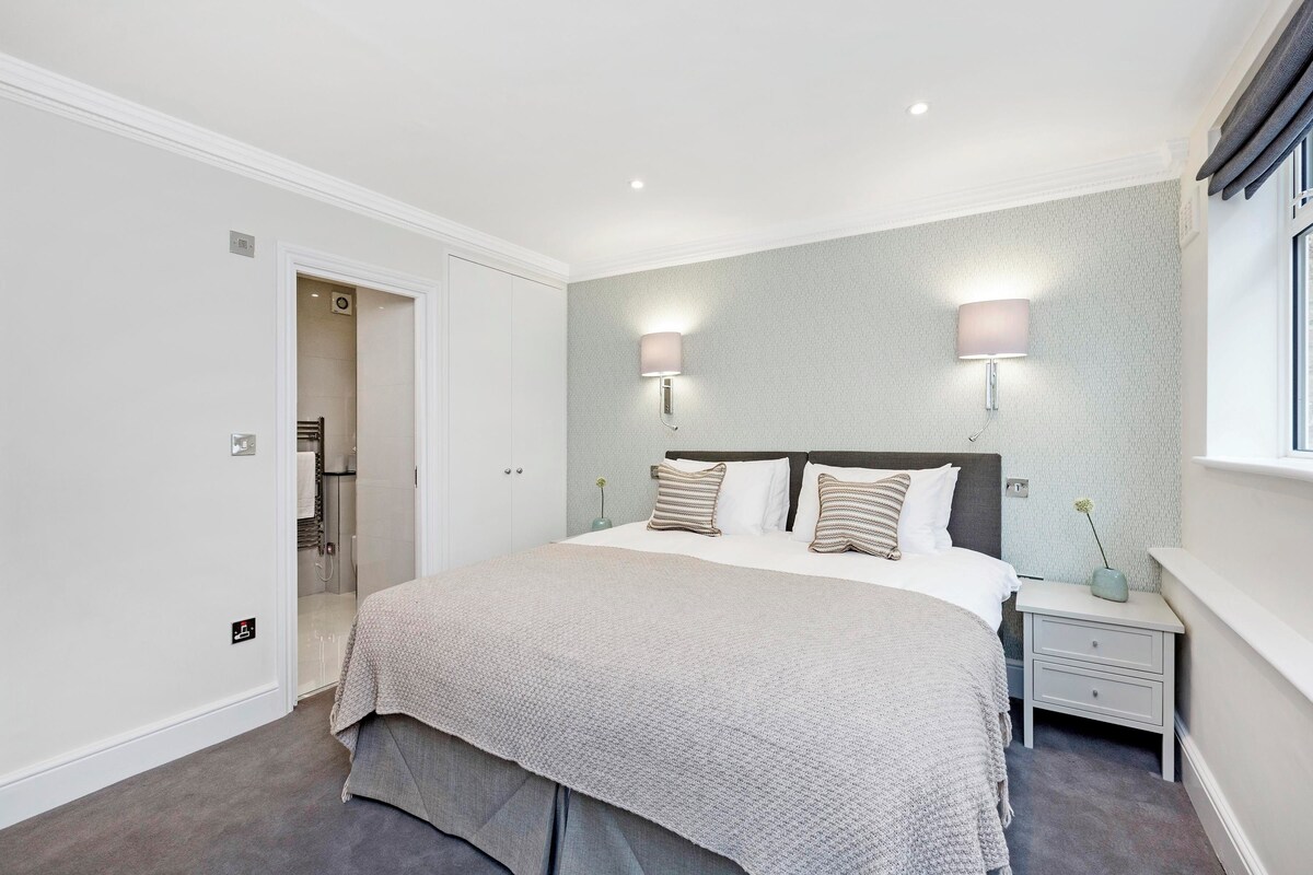 A bedroom with a large bed, two bedside tables, a nightstand, a window with curtains, and a door leading to a bathroom.