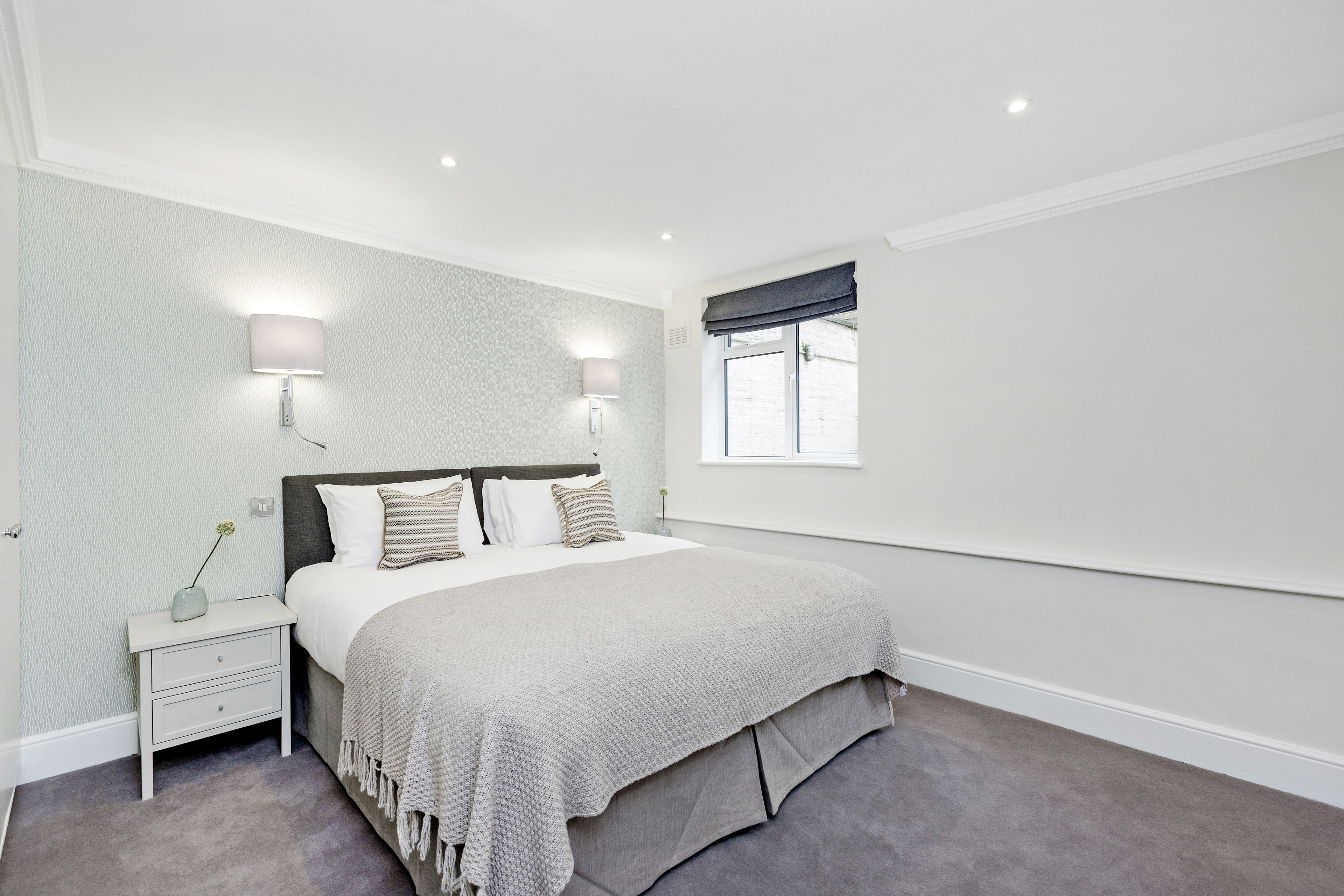 A bedroom with a bed, nightstand, wall-mounted lights, and a window.