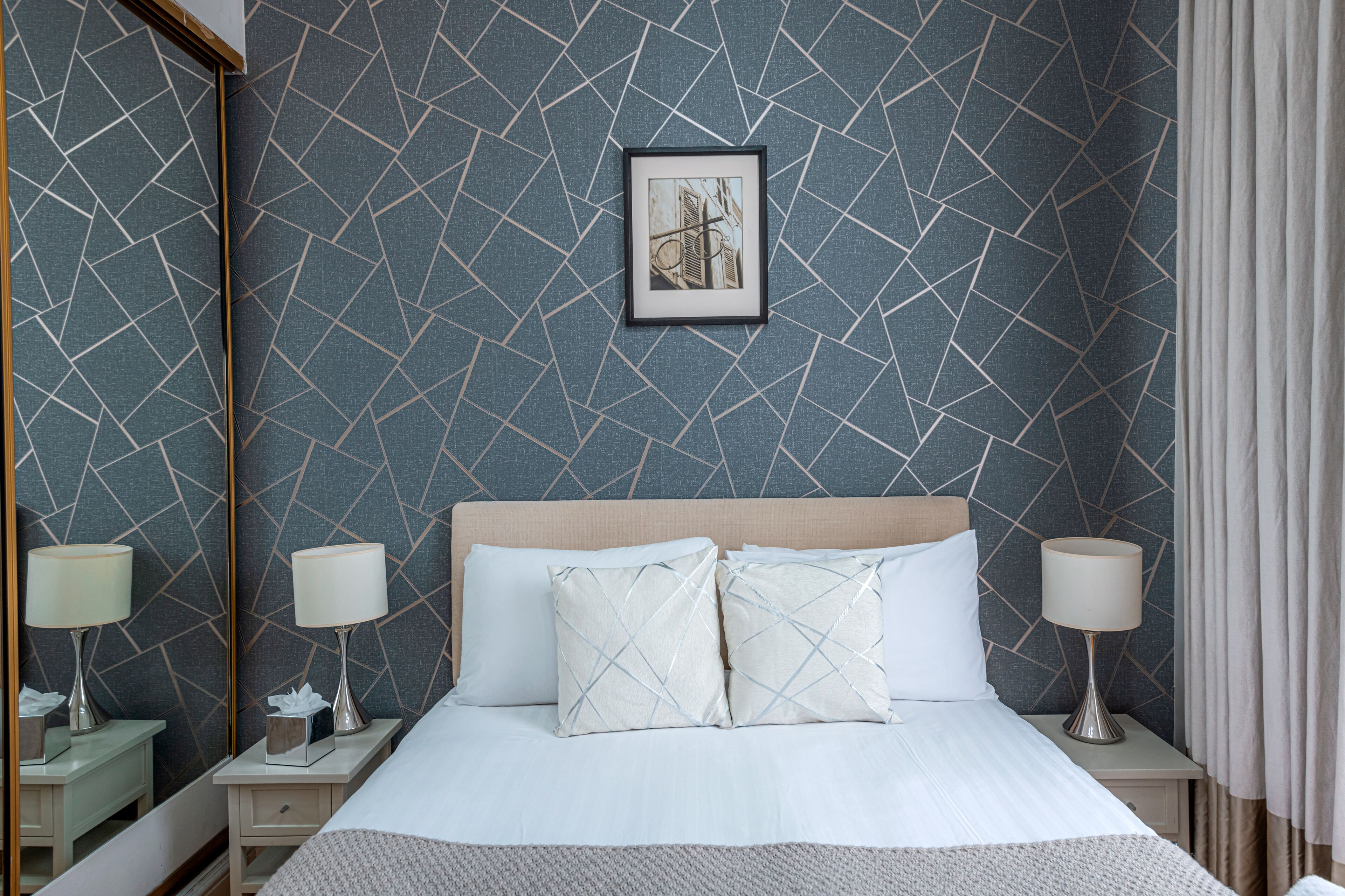 Una cama bien hecha con ropa de cama blanca y dos almohadones decorativos, flanqueada por mesitas de noche con lámparas y un cuadro en la pared.